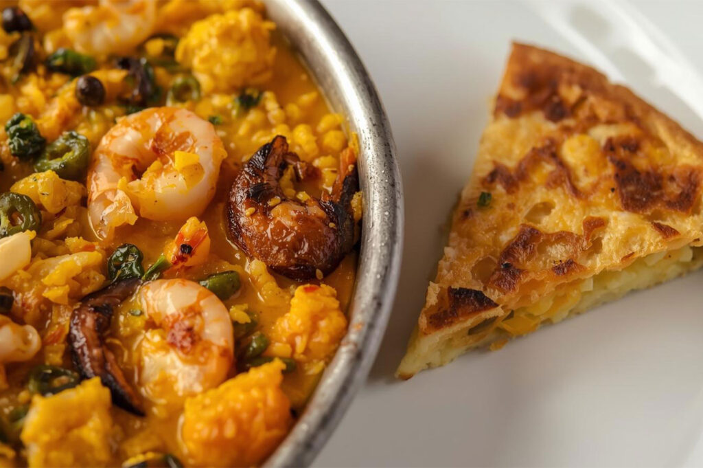 Close-up of a Spanish seafood paella with shrimp and saffron rice served in a metal pan, alongside a slice of traditional Spanish potato omelet (tortilla de patatas).