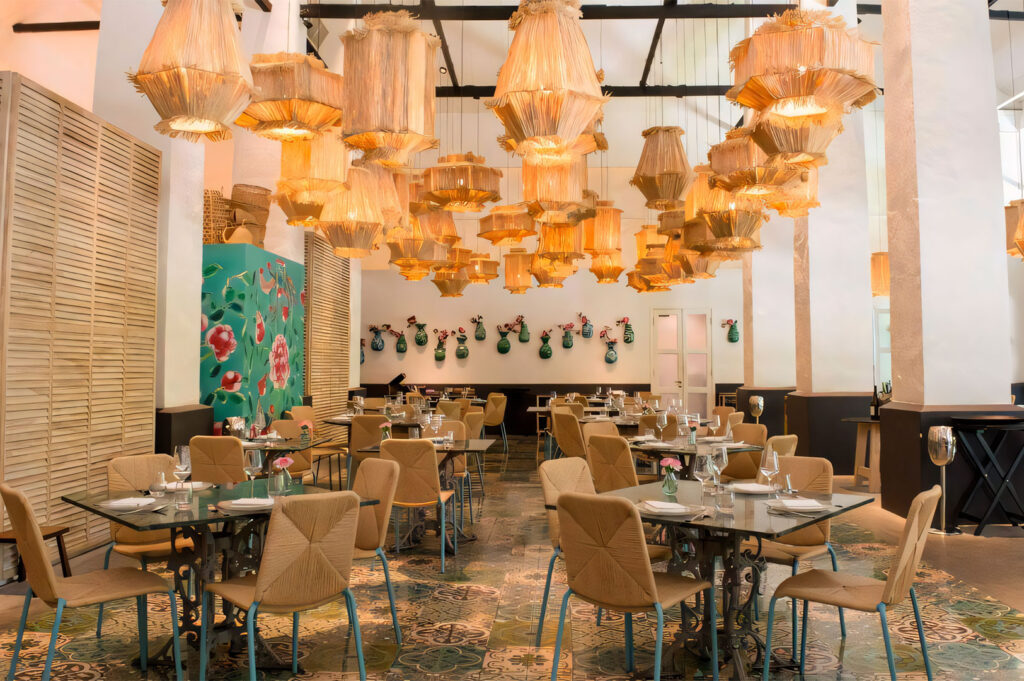 Spacious and brightly lit restaurant interior featuring an eclectic design with many large, woven wicker/straw basket chandeliers hanging from the ceiling. The dining area has small glass-top tables, modern chairs, and colorful patterned floor tiles.