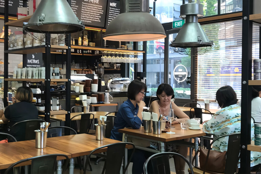 Three women sitting at a wooden table in a well-lit, stylish cafe with high shelves and industrial light fixtures, enjoying coffee and a conversation.