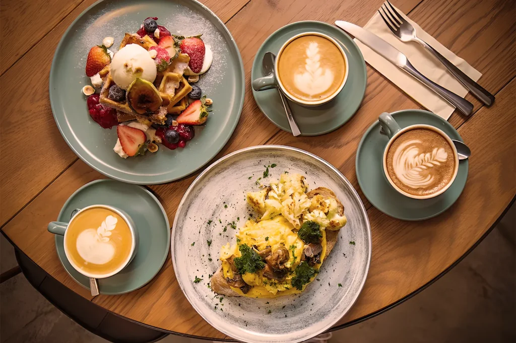 Gourmet brunch with waffles topped with berries and ice cream, scrambled eggs on toast, and artisan lattes with latte art.
