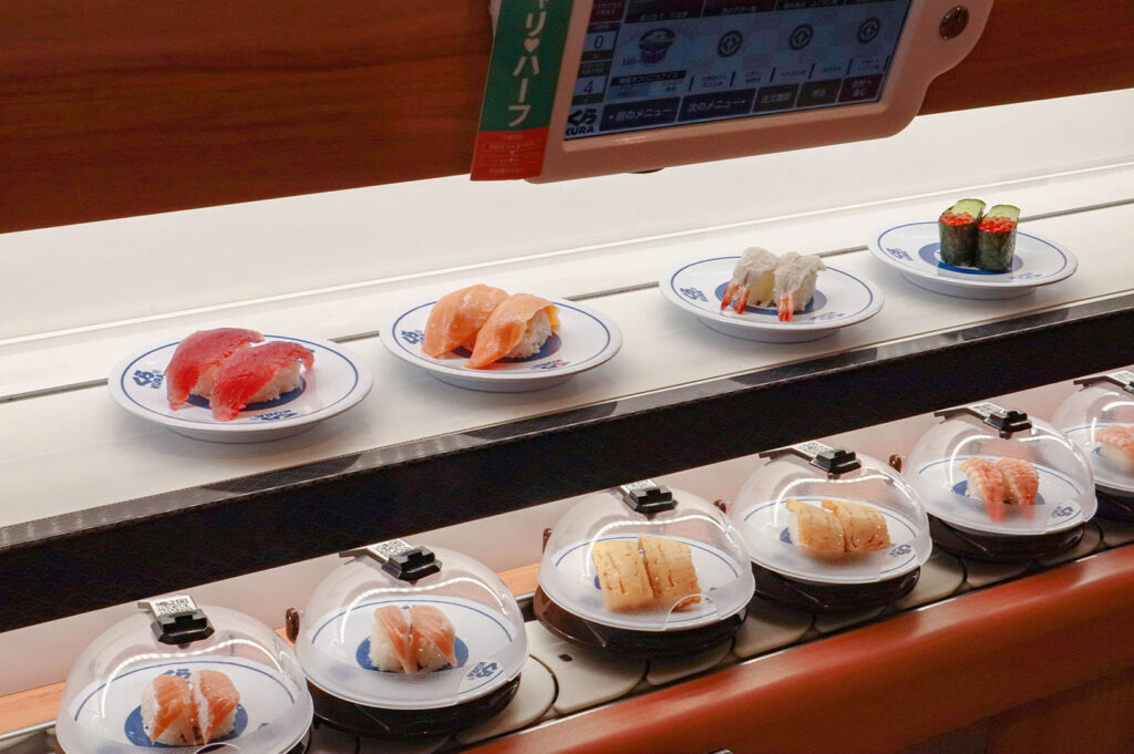 A brightly lit Japanese kaiten (conveyor belt) sushi restaurant, showing various plates of tuna, salmon, and tamago sushi moving along the belt, some covered by plastic domes, with a digital ordering screen visible above.