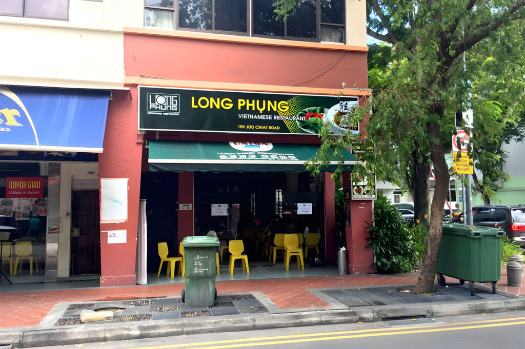 Exterior of Long Phung Vietnamese Restaurant on Joo Chiat Road, Singapore, featuring red walls, a green awning, and yellow plastic chairs on the sidewalk.