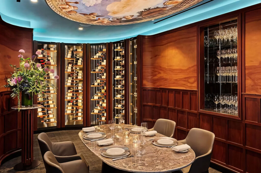 A luxurious, wood-paneled private dining room featuring a round marble table, leather chairs, a domed painted ceiling, and a prominent glass-fronted wine display cellar.