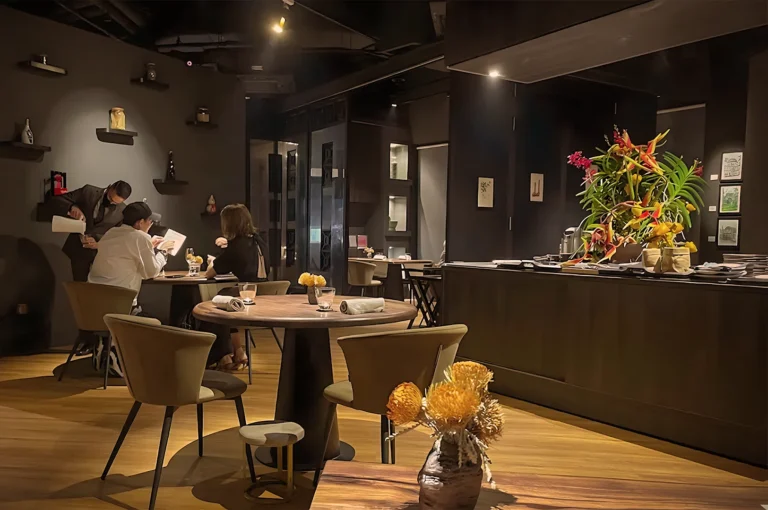 Modern, dimly lit restaurant interior with wooden floors. Three people at a table engage with menus. Bright floral arrangement on a counter adds warmth.