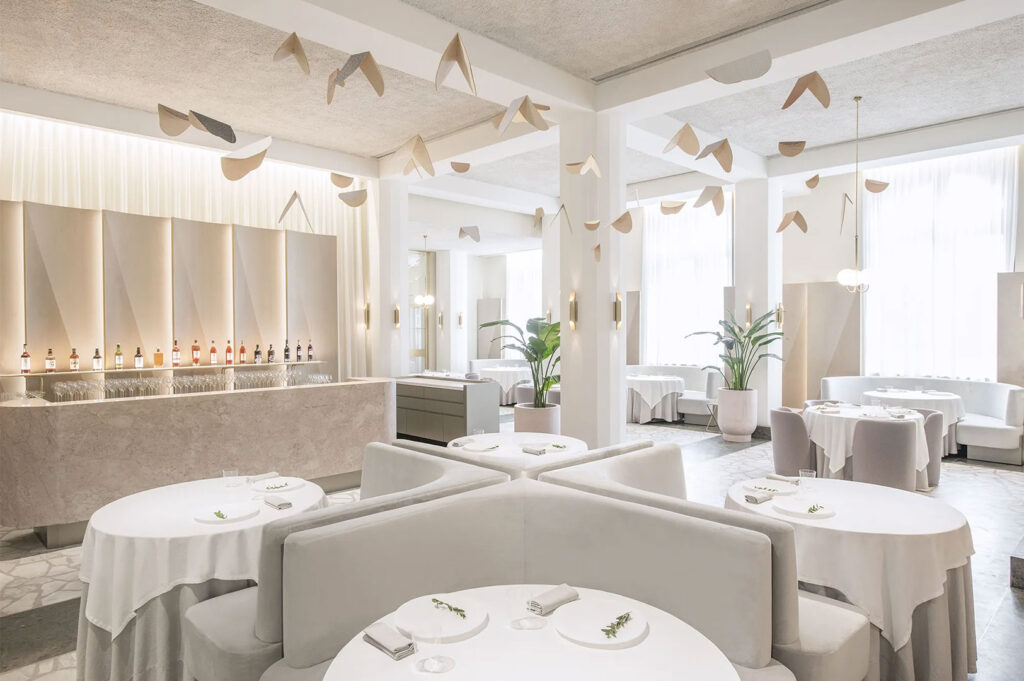 Bright, minimalist luxury restaurant interior with white and beige tones, curved velvet seating, round tables with white linens, and artistic paper mobiles hanging from the ceiling.