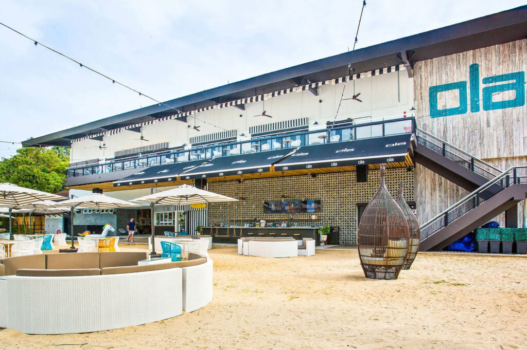 The exterior of the Ola Beach House on Sentosa Island, Singapore, showing a large building with a covered patio and stylish white and turquoise outdoor lounge seating on the sand.