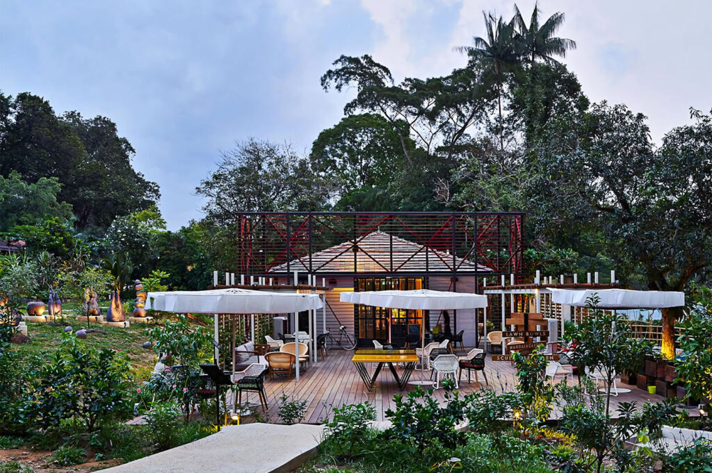 Exterior view of a charming, rustic outdoor restaurant and dining deck surrounded by lush greenery, trees, and gardens under an overcast sky.