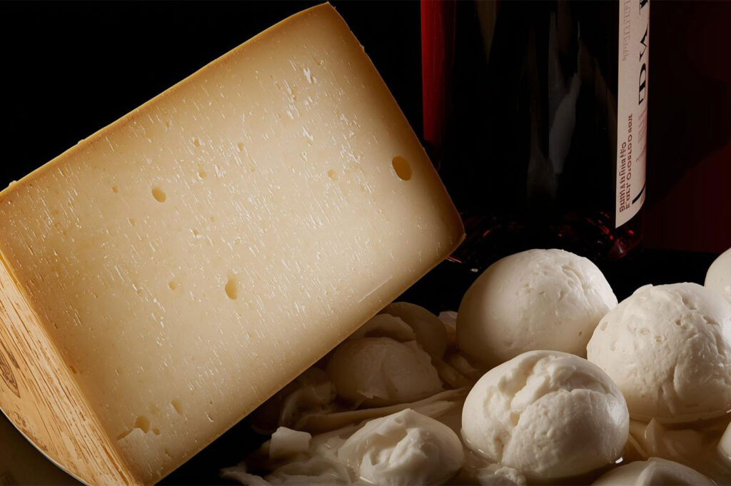 Close-up still life featuring a wedge of firm, aged Italian cheese (like Pecorino or Parmigiano) with characteristic holes, resting next to balls of fresh white mozzarella or burrata cheese, with a dark bottle of red wine visible in the background.