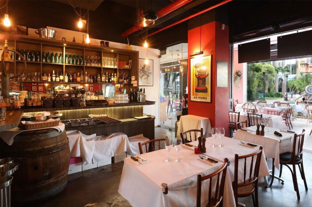 The cozy interior of a French-style bistro with a full bar and liquor shelf, rustic wooden barrel, tables set with white tablecloths, and a poster that reads "Air France" on a red wall.