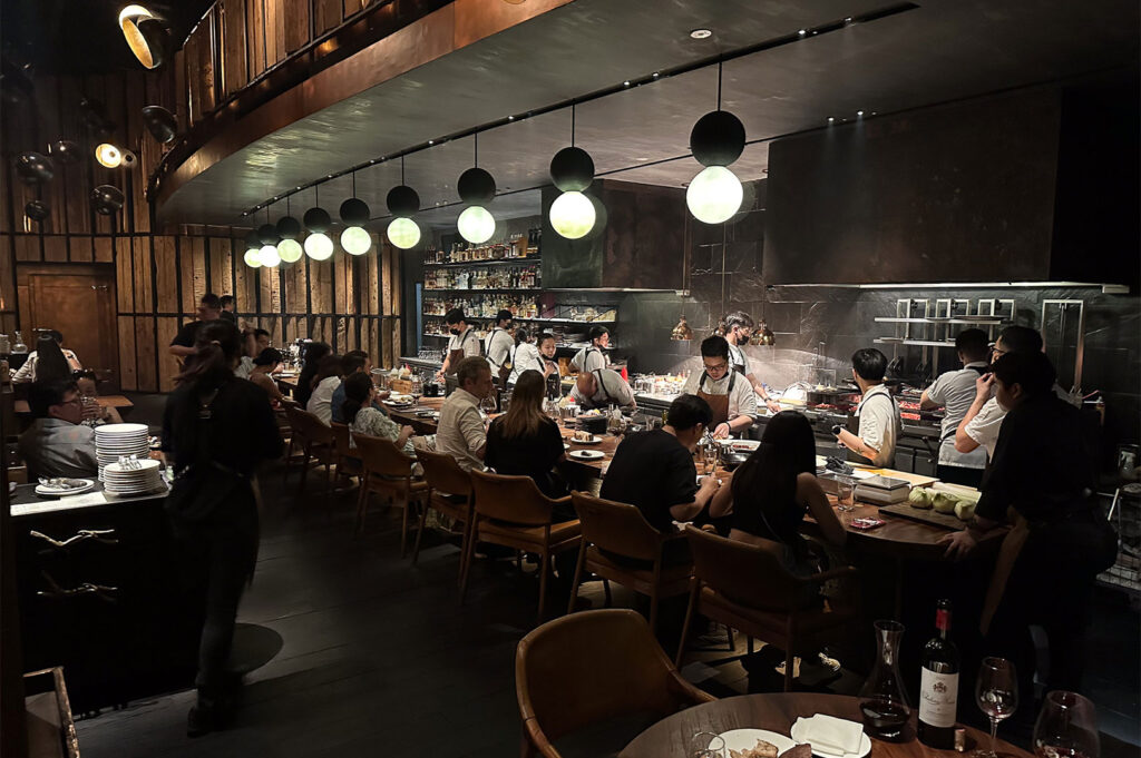 Darkly lit, modern restaurant interior showing patrons seated at a long counter bar facing an open kitchen. Several chefs are actively preparing food, illuminated by spherical pendant lights hanging over the bar area.