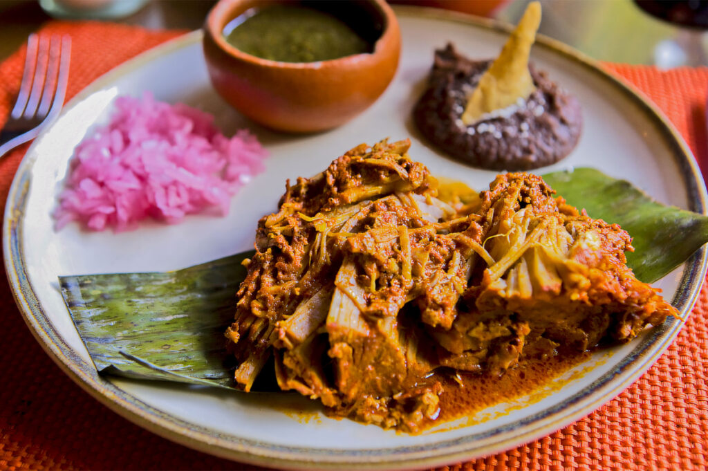 A close-up of a plate featuring a traditional Mexican dish, likely Cochinita Pibil (slow-roasted pork), resting on a banana leaf, served with a side of bright pink pickled red onions, refried beans, and a small terracotta bowl of salsa.