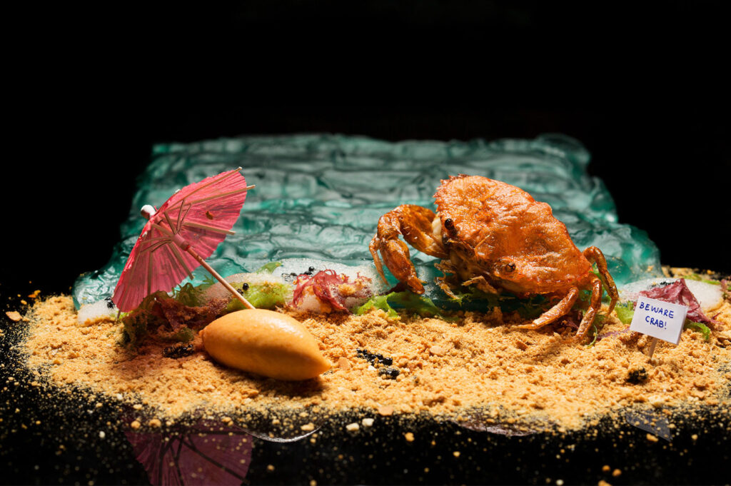 Highly artistic and conceptual chili crab dish presentation on a dark background, depicting a crab on 'sand' with a small pink umbrella and a sign saying "Beware Crab!"