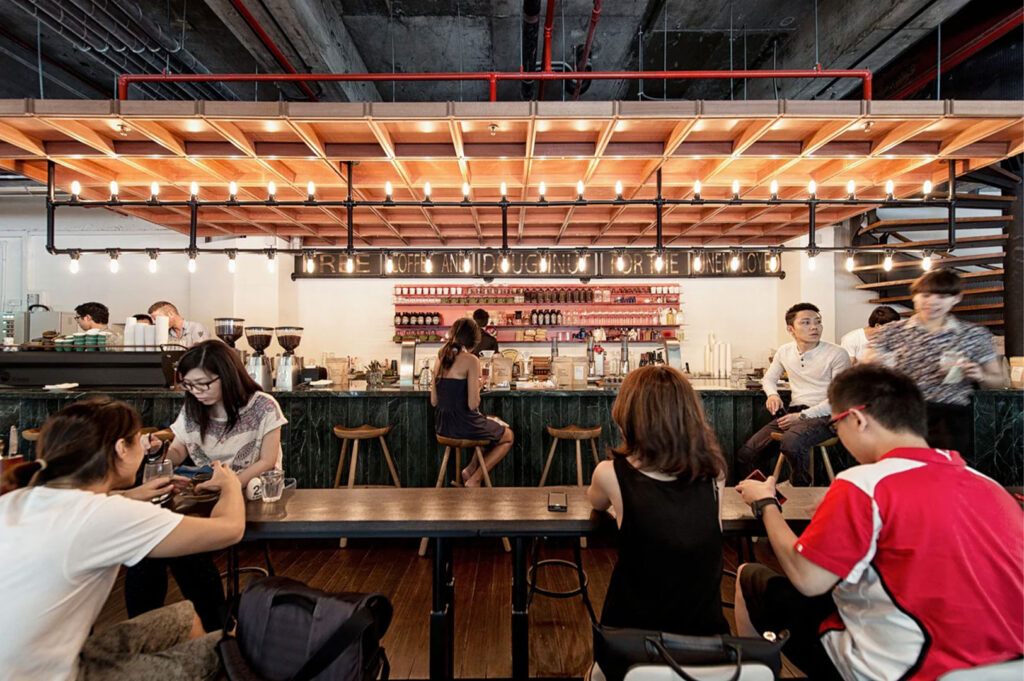 A wide shot of a bustling, trendy coffeeshop with industrial design, featuring a long communal table, a bar area, and customers chatting and using their phones.
