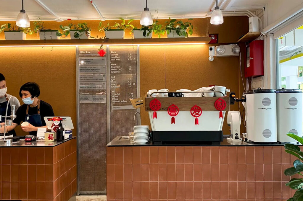 Coffee shop counter with exposed brick-colored tile, a large white espresso machine decorated with Chinese New Year ornaments, and a menu board detailing filter and espresso prices.