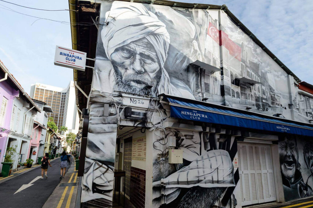Large black-and-white mural of an older man with a turban painted on the side of a building on Haji Lane, contrasting with the brightly colored shophouses nearby.