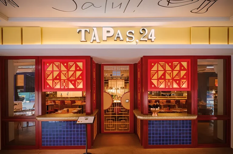 Entrance of a tapas bar named "TAPAS,24" with red geometric doors, blue tiles, and warm lighting, creating a welcoming and vibrant atmosphere.