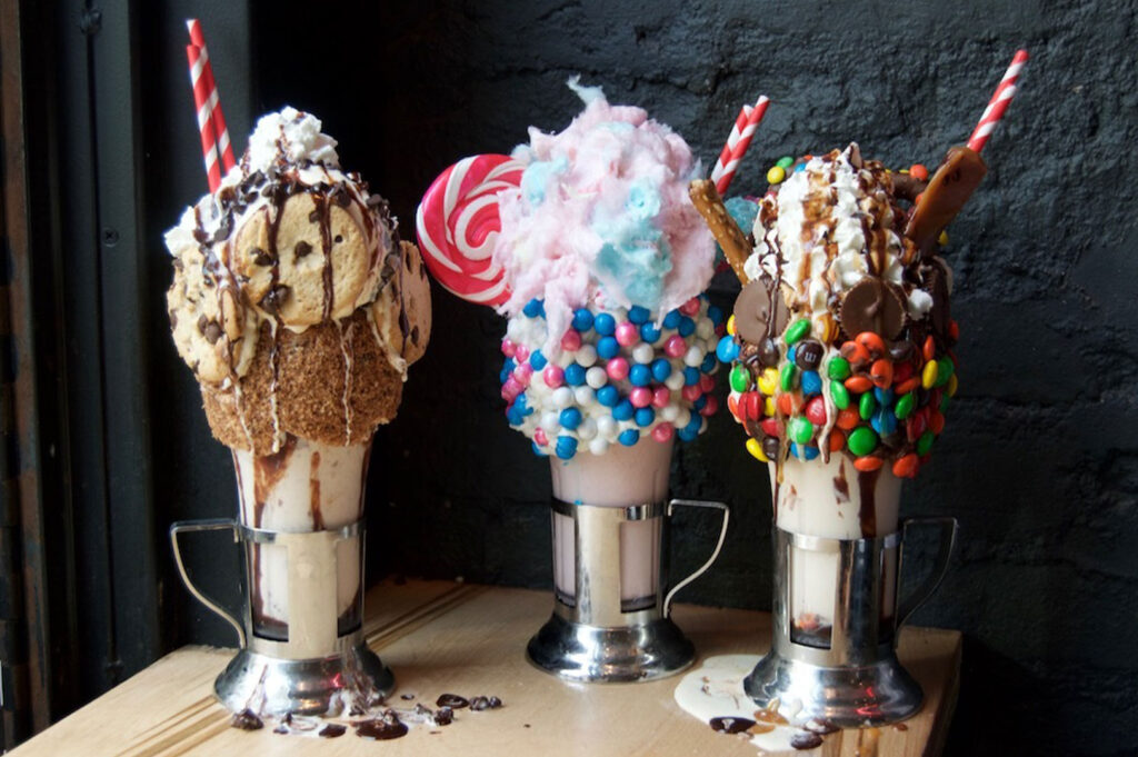 Three over-the-top "freakshakes" topped with whipped cream, cookies, M&Ms, lollipops, and cotton candy, served in metal milkshake glasses against a black background.