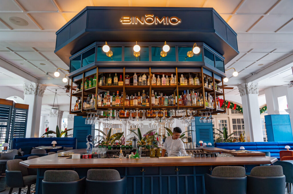 Stylish interior of a Spanish restaurant and bar, "Binomio," featuring a prominent navy blue hexagonal bar with a suspended liquor shelf and surrounding blue velvet seating.