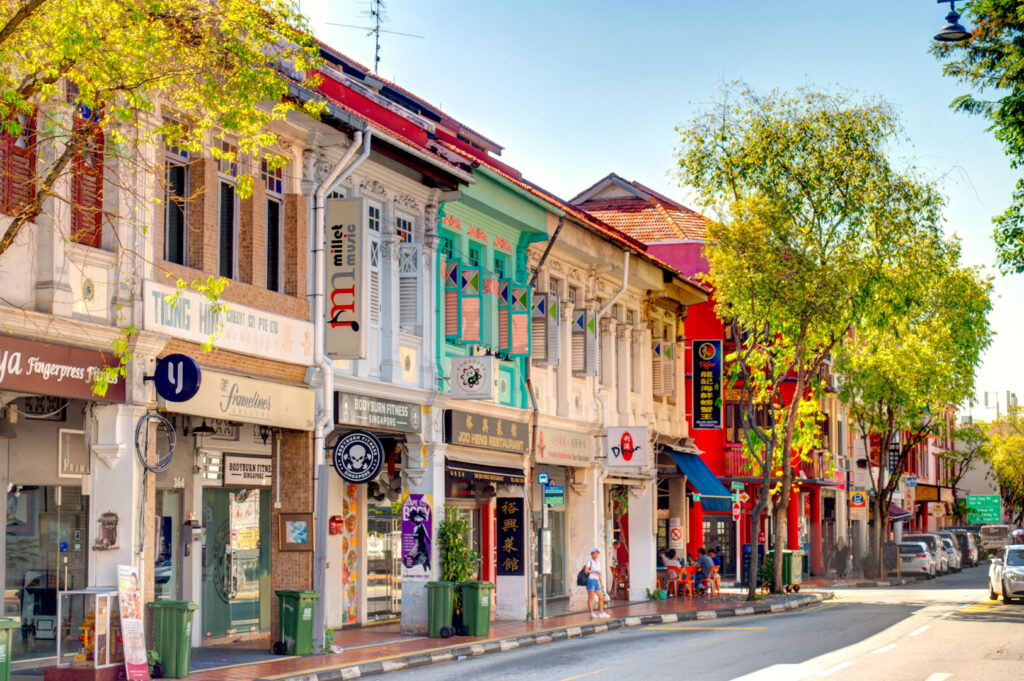 Sunny street view of a row of brightly colored, historic Peranakan shophouses along a street in Singapore (likely Joo Chiat or Katong), with various shops/businesses on the ground floor and lush green trees lining the street.