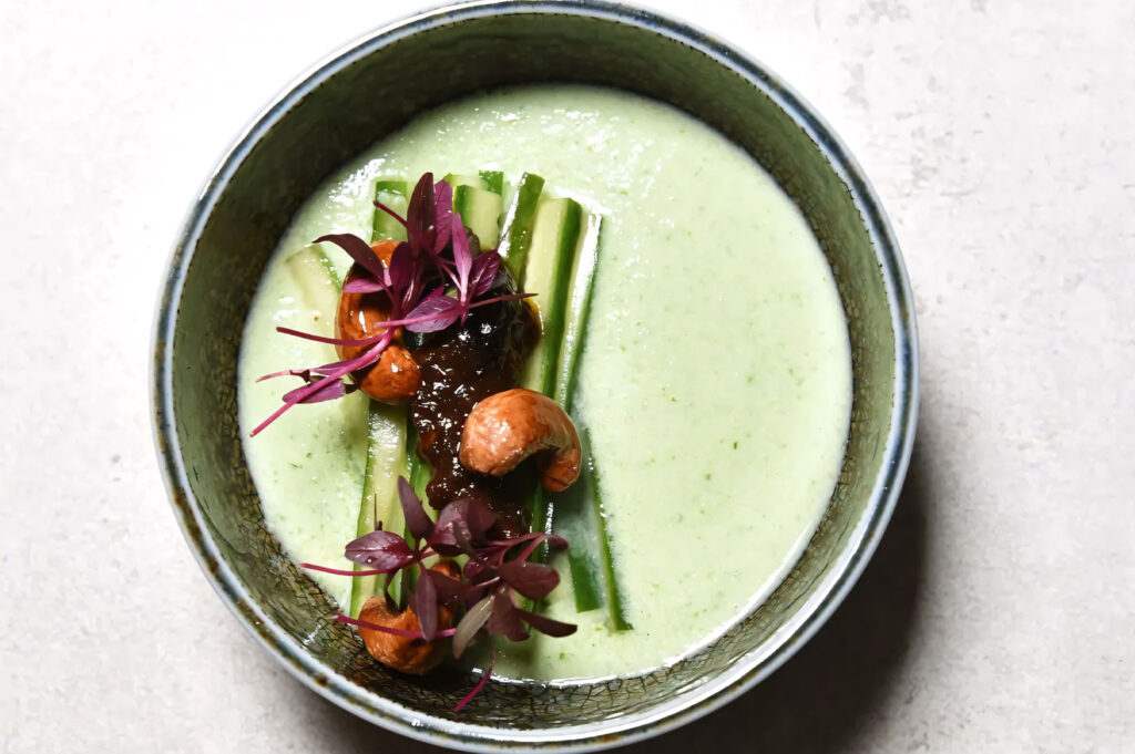 Overhead view of a fine dining green chilled soup or gazpacho in a ceramic bowl, garnished with cucumber strips, dark sauce, and purple microgreens.