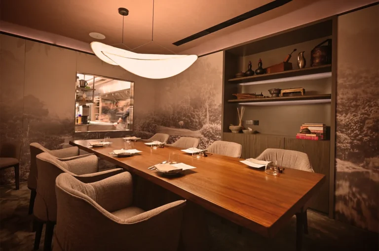 Elegant dining room with a long wooden table, eight cushioned chairs, soft lighting, and artistic wall decor. Open kitchen window shows a busy chef.