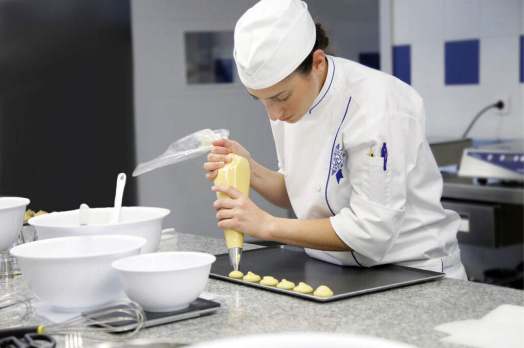 A focused female pastry chef in a white toque and coat using a piping bag to precisely deposit small rounds of dough (likely choux) onto a baking sheet in a professional kitchen setting.