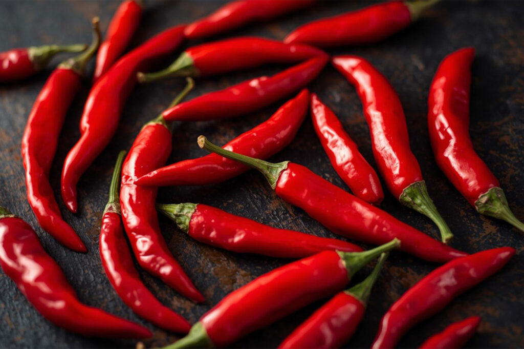 Overhead, close-up view of a scattered pile of long, bright red chili peppers with green stems, arranged on a dark, rustic background, emphasizing their color and texture.