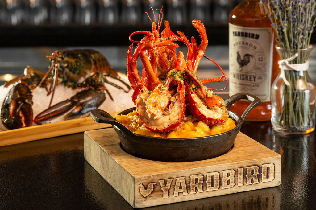 A black skillet of lobster mac and cheese from Yardbird, garnished with large lobster claws, next to a bottle of bourbon and a vase of lavender.