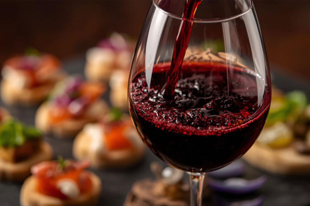 Close-up of deep red wine being poured into a stemmed glass, with a blurred background of various appetizers or canapés arranged on a dark platter.