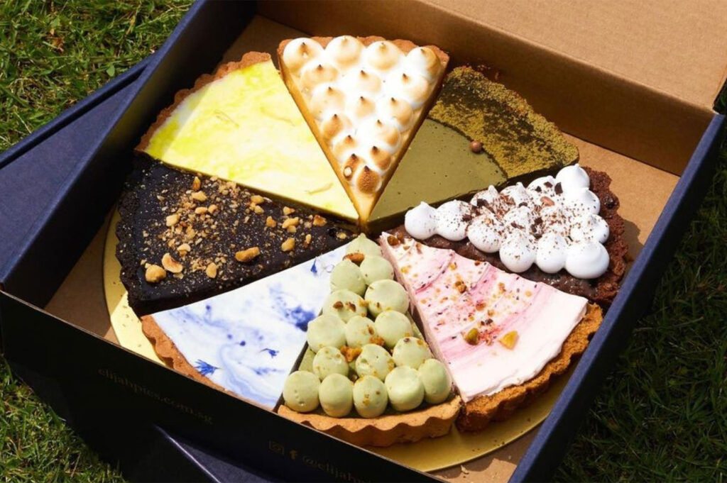 Close-up overhead shot of a sampler box from Elijah Pies, featuring eight slices of different flavored tarts and pies (e.g., lime, meringue, matcha, pistachio) set on a patch of green grass.