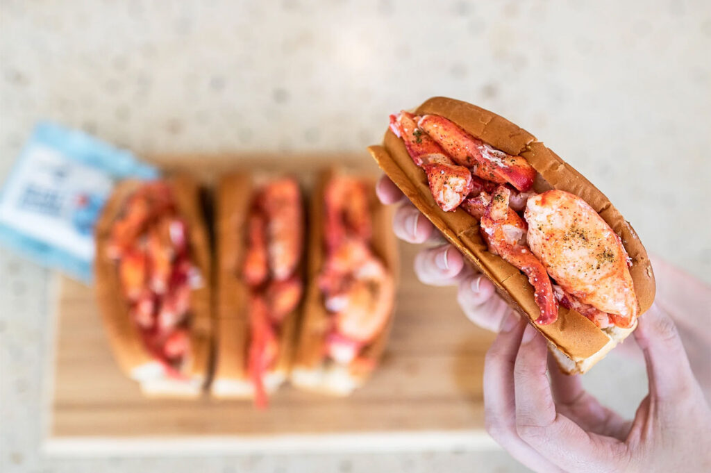 A close-up of hands holding a buttery New England-style lobster roll, with two more blurry lobster rolls and a wooden board in the background.
