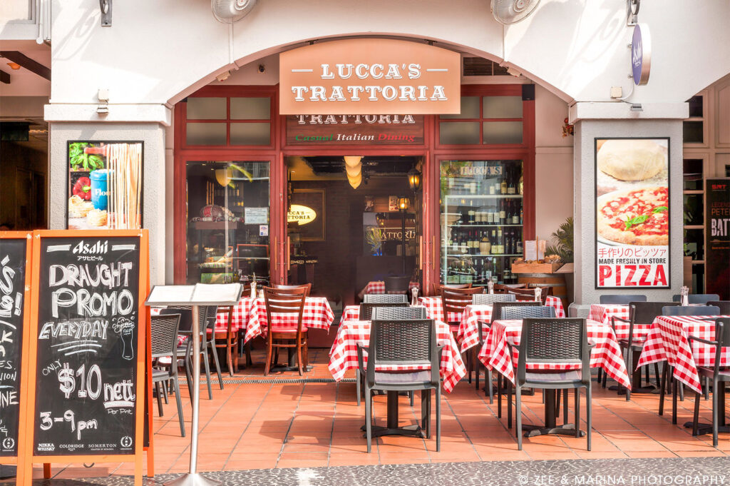 Exterior view of Lucca's Trattoria, an Italian restaurant with red-checkered tablecloths, outdoor seating, and a blackboard displaying a draught beer promo.