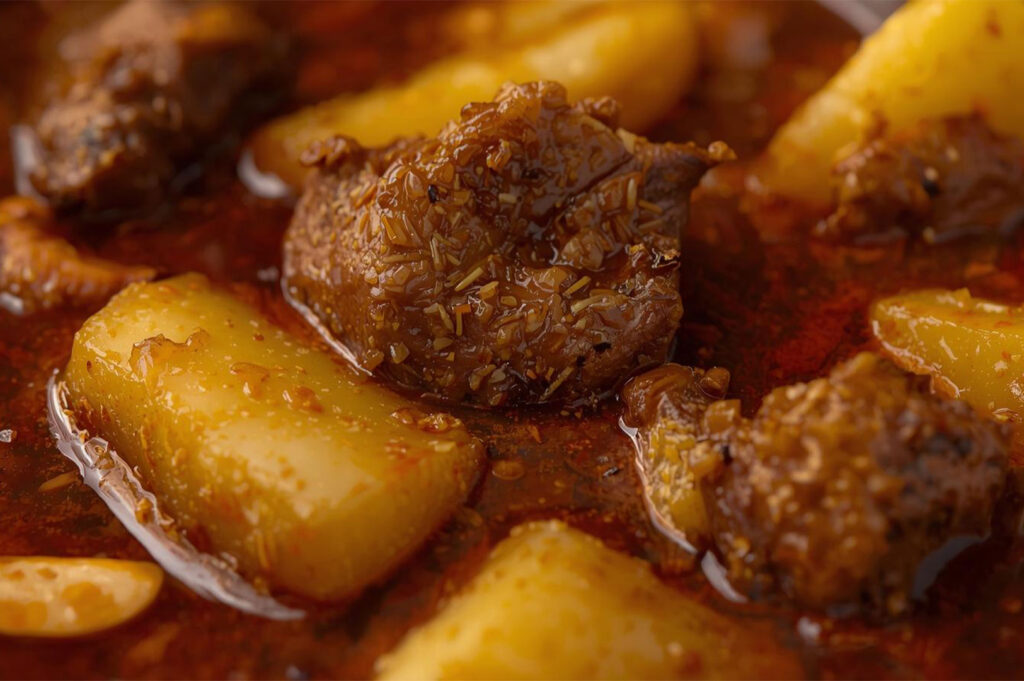 A macro close-up of a rich, dark red Southern Thai curry (likely Massaman or Panang), featuring chunks of beef or pork and large pieces of cooked yellow potato in a thick, oily sauce.