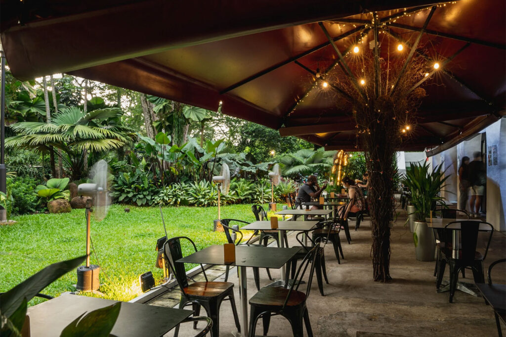 A shaded outdoor cafe patio area with black tables and chairs, looking out onto a vibrant green lawn and dense, lush tropical foliage, illuminated by string lights.