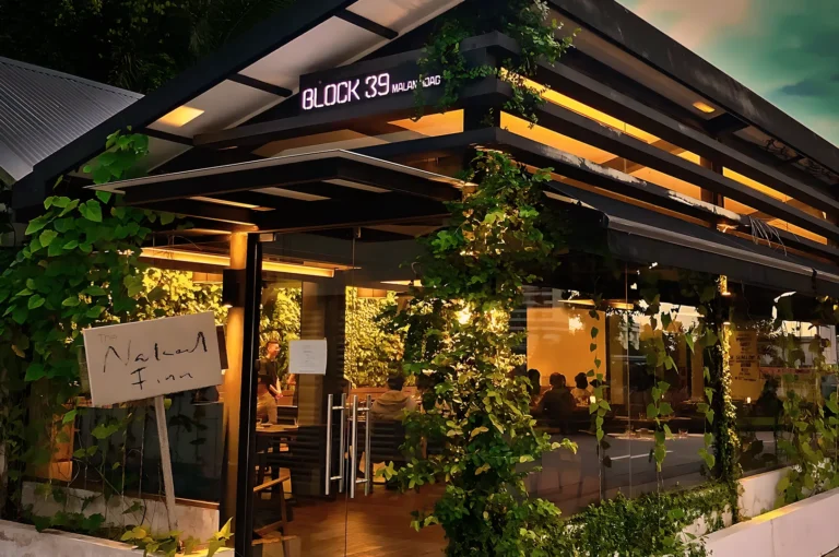 Exterior view of a modern café, "Block 39," at dusk. It has glass walls, warm lighting, and green vines climbing over the structure, creating a cozy vibe.
