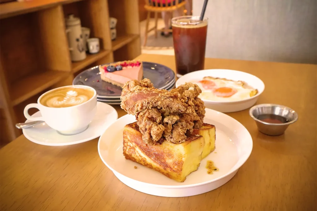 Café brunch featuring crispy fried chicken on French toast, latte art coffee, iced drink, and cheesecake dessert.