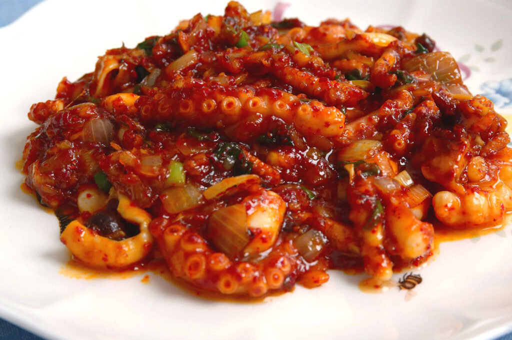 Close-up of a plate of vibrant, spicy Korean stir-fried octopus (likely Jjukkumi or Nakji Bokkeum) with onions and chili sauce.