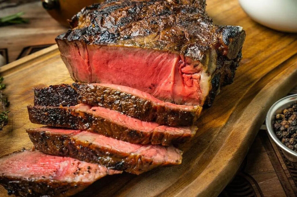 Close-up of a thick-cut, grilled prime rib steak, sliced to show a dark, charred crust and a perfect medium-rare pink interior, served on a wooden cutting board.