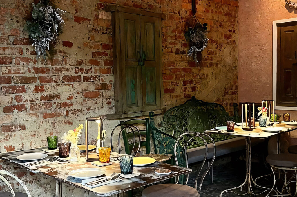 Rustic interior or patio of an Italian restaurant featuring an exposed brick wall, vintage distressed window shutters, and metal café seating.