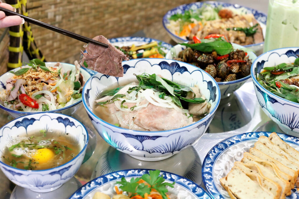 Close-up of a table laden with several bowls of Vietnamese dishes, including a central bowl of pho noodle soup from which chopsticks are lifting a slice of meat.