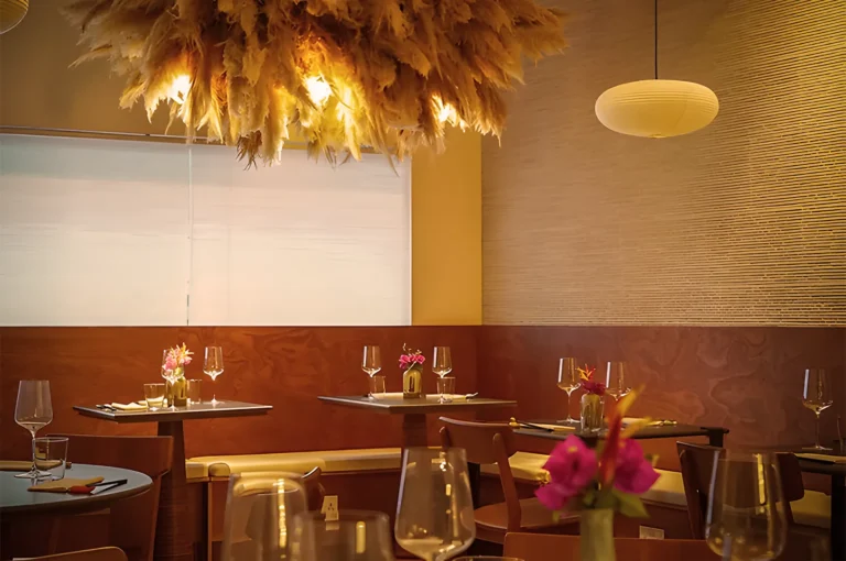 Cozy restaurant interior with warm lighting, pampas grass chandelier, and elegant table settings featuring pink flowers and sparkling glassware.