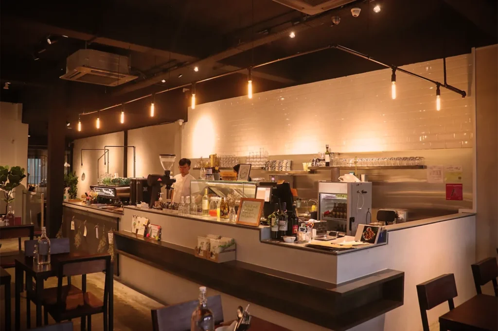 Warm modern café interior with barista at counter, hanging lights, espresso machine, and minimalist wooden tables.
