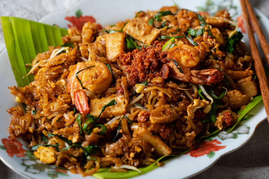 Close-up of a plate of Char Kway Teow, a dark, stir-fried rice noodle dish with shrimp, bean sprouts, tofu, and chives, served on a banana leaf liner.