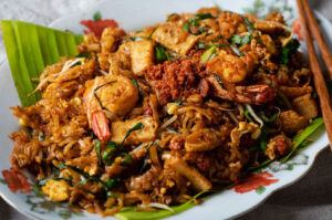 Close-up of a plate of Char Kway Teow, a dark, stir-fried rice noodle dish with shrimp, bean sprouts, tofu, and chives, served on a banana leaf liner.