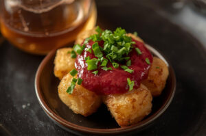 Close-up of crispy tater tots served in a small bowl, topped with a bright red chutney or house salsa and fresh chives, next to a glass of whiskey or cocktail