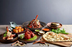 A lavish spread of Indian food on a wooden table. Key dishes include a centerpiece of lamb shank, grilled prawns, tandoori lamb chops, cottage cheese cubes, sauces, and a large piece of fresh naan bread.