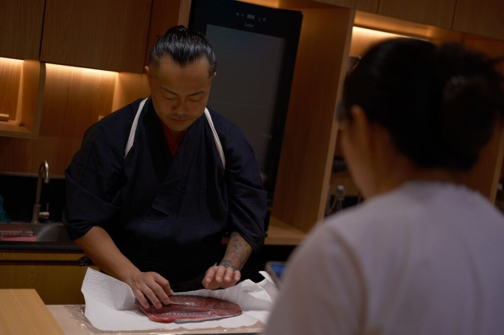 A man dressed as a chef cuts meat on a wooden board, showcasing culinary skills at Sushi Masa.
