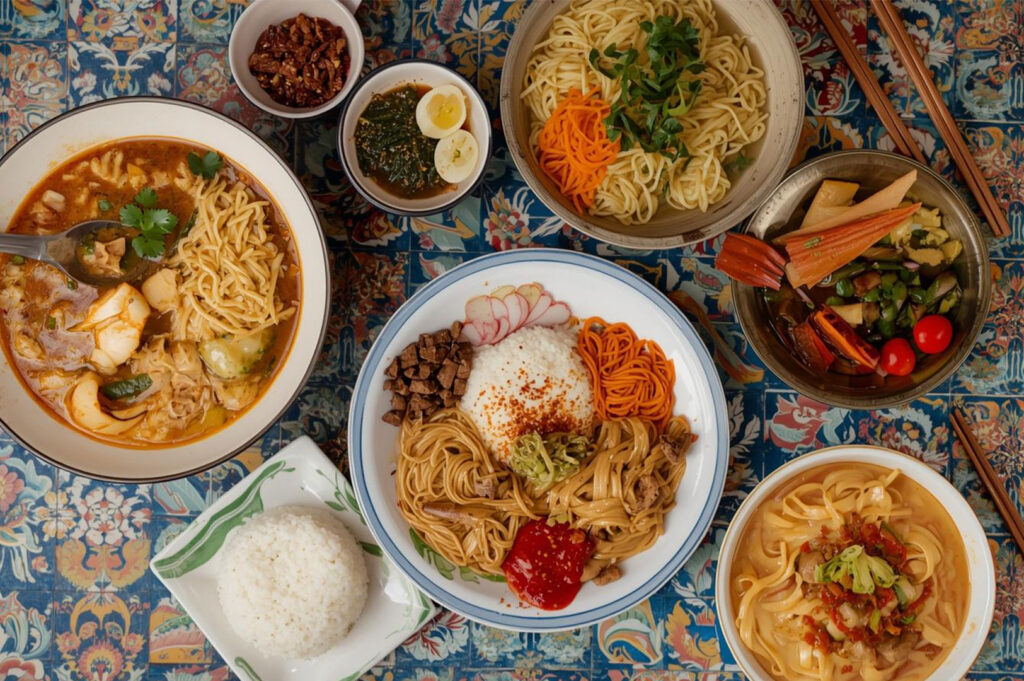 Overhead view of a vibrant table covered in a floral cloth, featuring various Asian noodle and rice dishes, including a spicy noodle soup, a dry noodle bowl with rice, and side condiments.