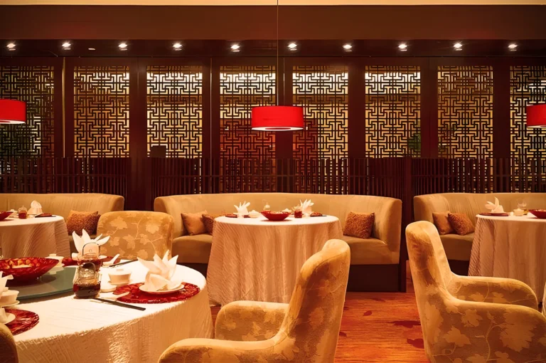 Luxurious Chinese restaurant interior with round tables, plush beige chairs, red pendant lights, and intricate lattice wall panels — refined fine dining atmosphere.