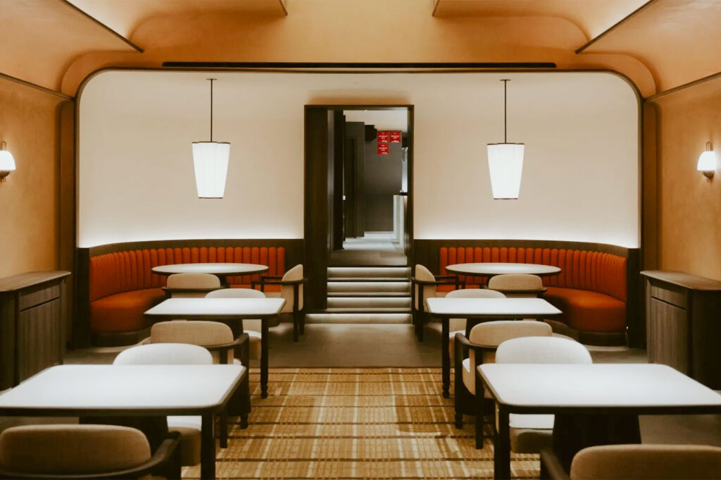 A view of a fine dining restaurant interior with a warm, modern aesthetic. Features two rows of tables, plush orange booth seating against the walls, and contemporary pendant lights hanging over the central aisle.